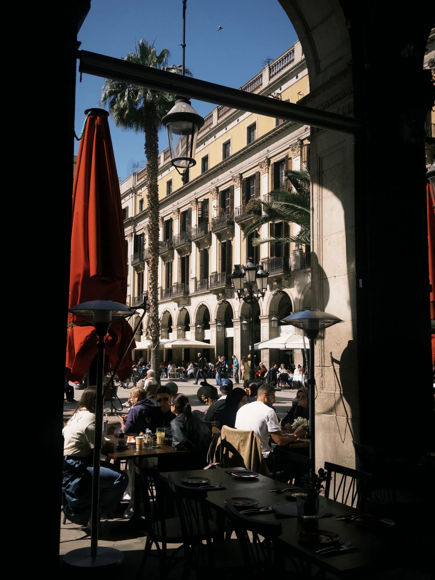 Terraza de café con sombrillas rojas junto a una plaza porticada, al atardecer.