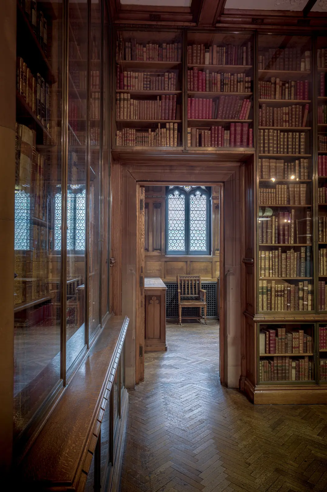 Pasillo de una biblioteca histórica con estanterías de libros antiguos y una vidriera al fondo.